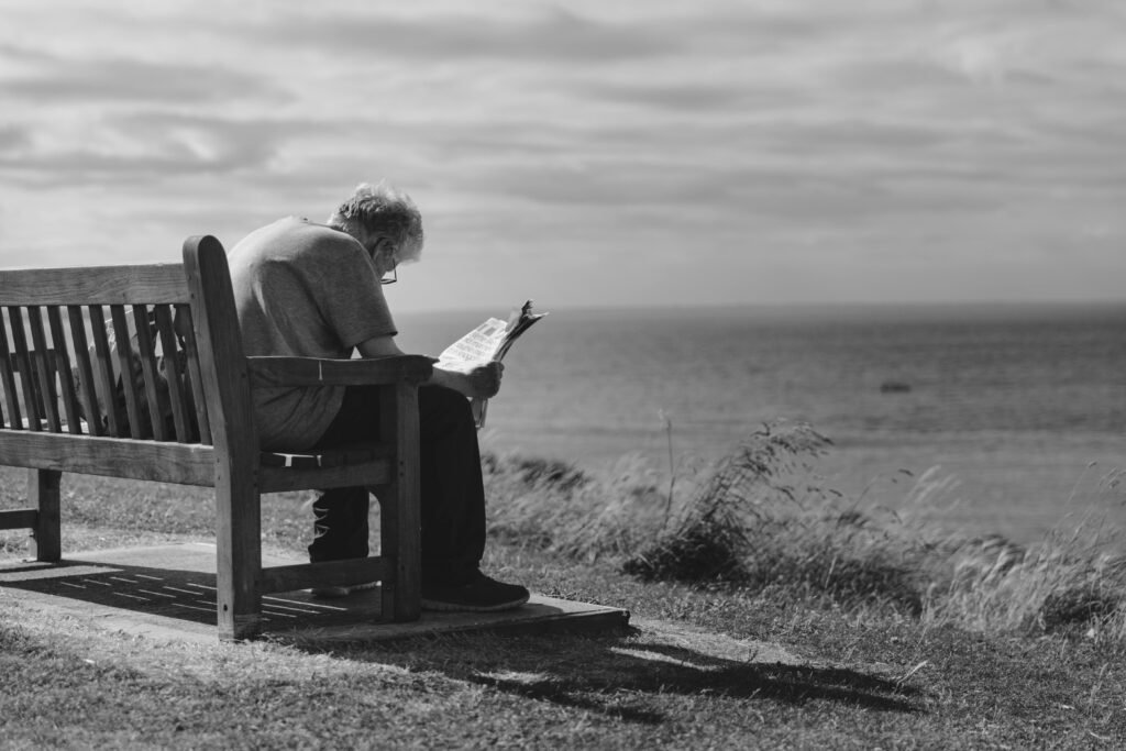 A contemplative scene of an elderly man reading a newspaper on a seaside bench with cloudy skies.